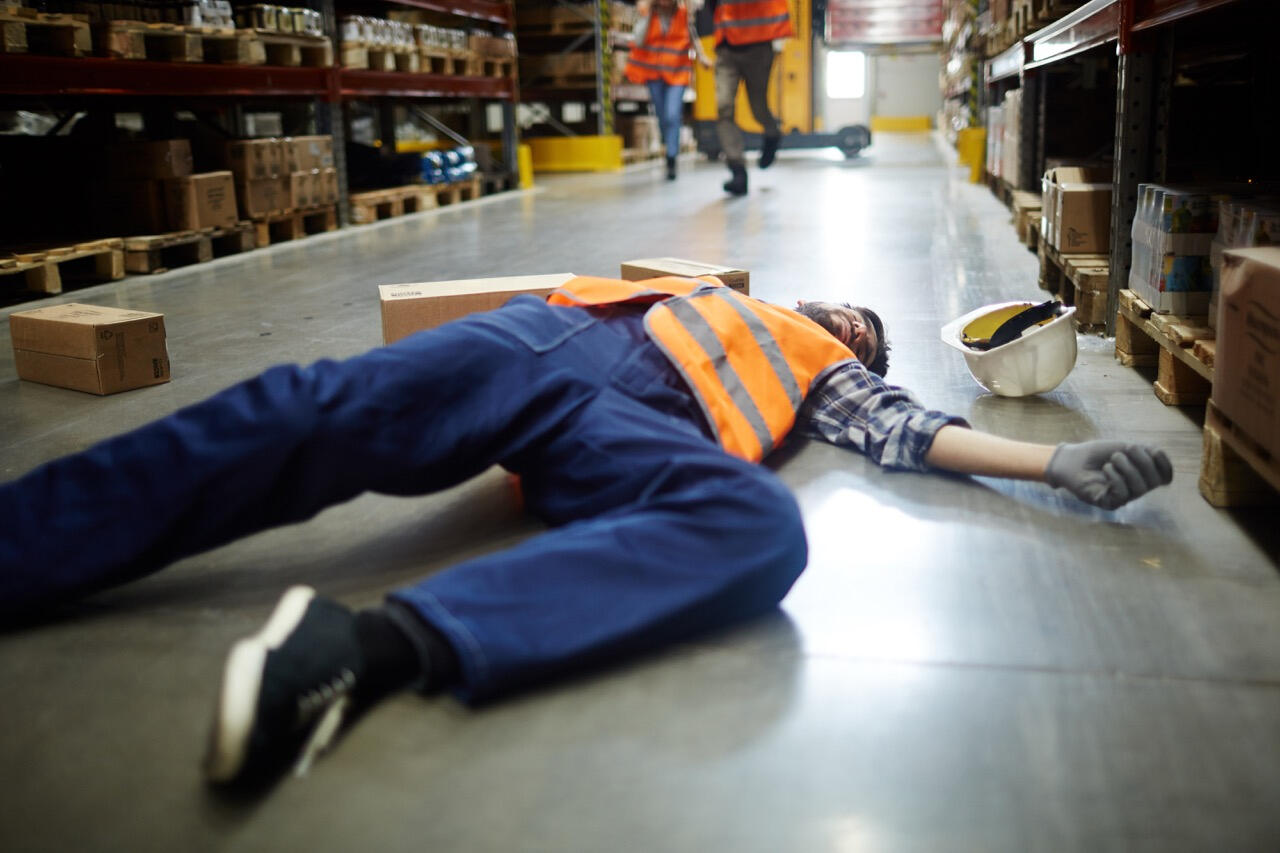 Workers Compensation Claim Investigation in a Warehouse Setting Wide-angle view of a warehouse worker in a safety vest lying unconscious on the floor surrounded by fallen boxes, illustrating workers compensation fraud detection services by West Coast Private Investigators in Rancho Santa Margarita, CA